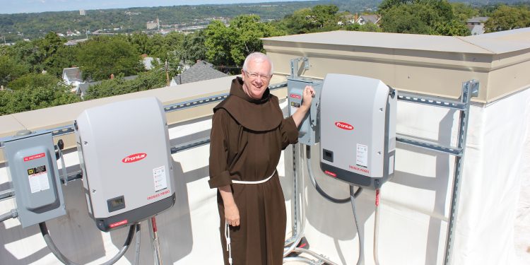 Franciscan Father Al Hirt, pastor of St. Monica-St. George Parish Newman Center, prepares to throw the switch on the inverters that convert solar power DC to alternating current on the roof of the parish center recently. (Courtesy Photo)