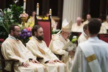 Archbishop Robert G. Casey sits beside two deacons and preaches his homily.