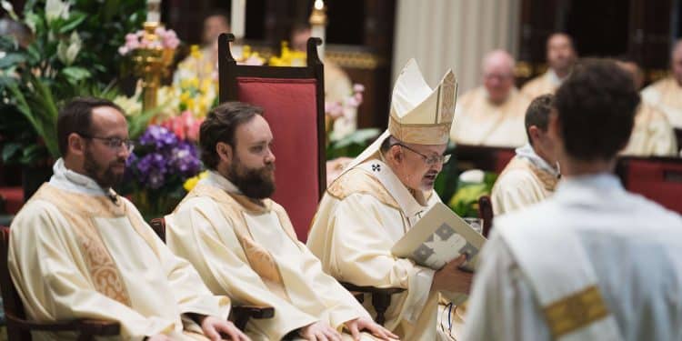 Archbishop Robert G. Casey sits beside two deacons and preaches his homily.